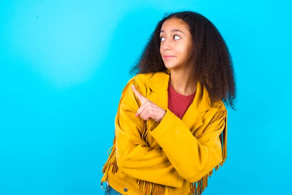青い背景に黄色のジャケットを着たアフロスタイルのアフリカ系アメリカ人のティーンエイジャーの少女の肖像画は トリッキーな外観でカメラにポーズをとって インデックスの指で製品を提示します 広告コンセプト — ストック写真