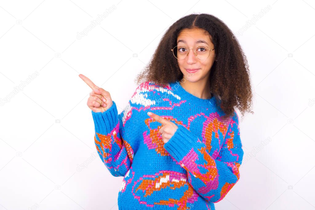 African American teenager girl glasses  with afro hair style wearing vintage colourful sweater over white background points at copy space indicates for advertising gives right direction
