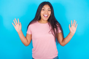İyimser kadın sevinçten avuçlarını kaldırır, birinden müthiş bir hediye aldığı için mutludur, yüksek sesle bağırır, heyecanlı bir manken gibi çığlık atar. Pembe tişört giyen esmer, güzel bir kadın mavi arka plana poz veriyor.