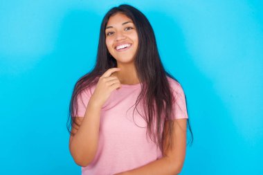 İyimser kadın ellerini kısmen çapraz yapar ve elleri çenesinin altında tutar, kameraya zevkle bakar. Mutlu duygular kavramı. Pembe tişört giyen esmer, güzel bir kadın mavi arka plana poz veriyor.