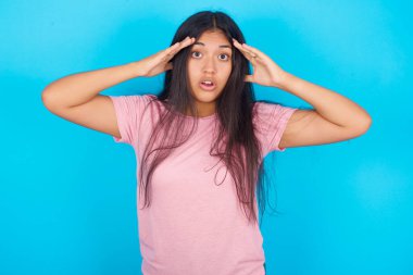 Yüzünde korku ifadesi olan genç bir kız, elleri başının üstünde, çenesi düşmüş, müthiş bir ifadesi var. Omg konsepti. Pembe tişört giyen esmer, güzel bir kadın mavi arka plana poz veriyor.