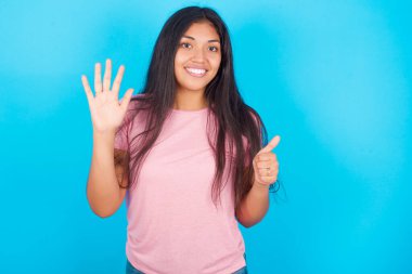 Genç kız kendini beğenmiş ve mutlu bir şekilde gülümserken altı numarayı işaret ediyor. Pembe tişört giyen esmer, güzel bir kadın mavi arka plana poz veriyor.