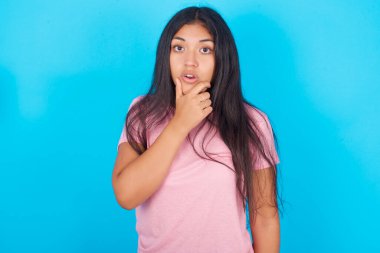 İnançsızlıktan, şaşkınlıktan ve elleri çenesinde olan şaşkınlıktan büyülenmiş genç bir kız. Pembe tişört giyen esmer, güzel bir kadın mavi arka plana poz veriyor.