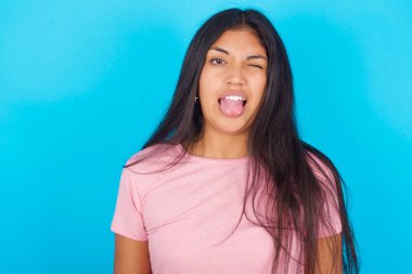 Genç kız komik bir ifadeyle dilini dışarı çıkartıyor. Duygu konsepti. Pembe tişört giyen esmer, güzel bir kadın mavi arka plana poz veriyor.