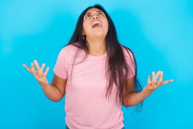 Genç kız deli gibi bağırıyor, bağırıyor ve agresif bir ifadeyle kollarını kaldırıyor. Hayal kırıklığı konsepti. Pembe tişört giyen esmer, güzel bir kadın mavi arka plana poz veriyor.