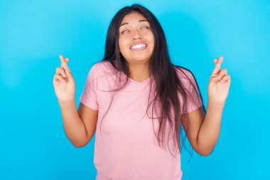 Umudu ve gözleri kapalı gülümseyen işaret eden genç bir kız. Şans ve batıl inançlar. Pembe tişört giyen esmer, güzel bir kadın mavi arka plana poz veriyor.