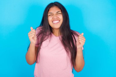 Genç kız zafer kutlamaları için kollarını kaldırarak ve gözlerini kapatarak başarı için heyecanlanıyor. Kazanan konsept. Pembe tişört giyen esmer, güzel bir kadın mavi arka plana poz veriyor.