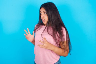  Kız yüksek sesle bağırıyor, gözleri açık ve elleri gergin tutuyor. Pembe tişört giyen esmer, güzel bir kadın mavi arka plana poz veriyor.