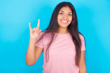  Kaya hareketi yapan ve kameraya gülümseyen bir kız. En sevdiği konsere gitmeye hazır. Pembe tişört giyen esmer, güzel bir kadın mavi arka plana poz veriyor.