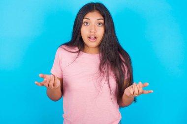 Hayal kırıklığına uğramış kız şaşkın ve tereddütlü hissediyor, şaşkınlıkla omuz silkiyor, ağzını sonuna kadar açık tutuyor, ne yapacağını bilmiyor. Pembe tişört giyen esmer, güzel bir kadın mavi arka plana poz veriyor.