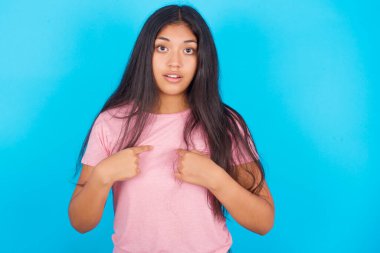 Utangaç bir kız, şaşkın bir ifadeyle yarışmaya katıldığı için şoka uğruyor, bir konuda tereddüt ediyor. Pembe tişört giyen esmer, güzel bir kadın mavi arka plana poz veriyor.