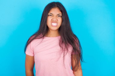  Kız dişlerini sıkar, kaşlarını çatar, tatminsizlikten suratını asar, görevleri yüzünden tedirgin olur. Pembe tişört giyen esmer, güzel bir kadın mavi arka plana poz veriyor.