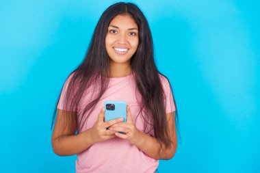  Kız uzak iletişimden hoşlanıyor, cep telefonu kullanıyor, hızlı limitsiz internet sörfü yapıyor, hoş bir gülümsemesi var, online alışveriş yapıyor. Pembe tişört giyen esmer, güzel bir kadın mavi arka plana poz veriyor.