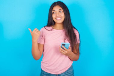  Kız akıllı telefonuyla mesaj atıyor ve gülümseyen yüzüyle baş parmağıyla işaret ediyor. Pembe tişört giyen esmer, güzel bir kadın mavi arka plana poz veriyor.
