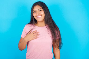  Kız dişleriyle gülümsüyor. Gözlerinin iyi duyguları ve şaşkınlığı ifade ettiğine inanamıyor. Pembe tişört giyen esmer, güzel bir kadın mavi arka plana poz veriyor.