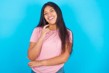  Kız mutlu bir şekilde gülüyor. Elini çenesine koyuyor. Olumlu duygular ifade ediyor. Pembe tişört giyen esmer, güzel bir kadın mavi arka plana poz veriyor.