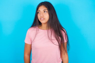  Kız şaşırmış bir ifadeyle yan yana bakıyor. Dili tutulmuş bir ifade var. Utangaç model şaşırmış görünüyor. Pembe tişört giyen esmer, güzel bir kadın mavi arka plana poz veriyor.