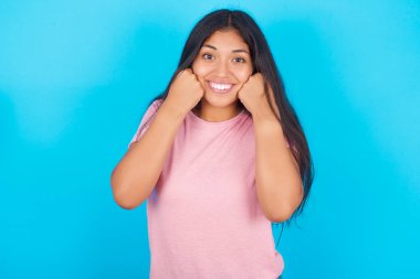 Mutlu kız yumruklarını yanaklarına dayayıp gülümsüyor ve morali yerinde olduğu için olumlu bir ifade takınıyor. Pembe tişört giyen esmer, güzel bir kadın mavi arka plana poz veriyor.