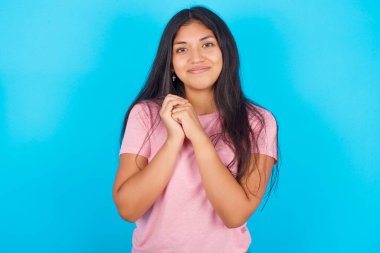 Çekici, ciddi bir kız kameraya nazikçe gülümseyen yüzler takınır. Pembe tişört giyen esmer, güzel bir kadın mavi arka plana poz veriyor.