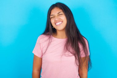 Neşeli yüzü olan pozitif kadın gözlerini kapar ve kahkahalar beyaz mükemmel dişleri gösterir. Pembe tişört giyen esmer, güzel bir kadın mavi arka plana poz veriyor.