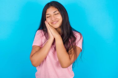  Kadın avuçlarına yaslanır, gözlerini kapatır ve bir şey hakkında hoş bir gülümseme görür. Pembe tişört giyen esmer, güzel bir kadın mavi arka plana poz veriyor.