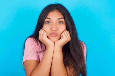  Şaşırmış yüz ifadesi olan bir kadın ellerini çenesinin altında tutar dudaklarını katlayarak suratını buruşturur. Pembe tişört giyen esmer, güzel bir kadın mavi arka plana poz veriyor.