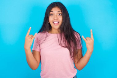  Kadın rock 'n roll tabelasını kendinden emin ve neşeli gösteriyor. Partide havalı müzikten hoşlanıyor. Vücut dili kavramı. Pembe tişört giyen esmer, güzel bir kadın mavi arka plana poz veriyor.