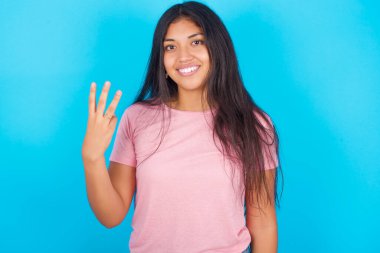  Kadın gülümsüyor ve arkadaş canlısı görünüyor. Üç ya da üç numarayı gösteriyor. Elleri önde, geriye doğru sayıyor. Pembe tişört giyen esmer, güzel bir kadın mavi arka plana poz veriyor.