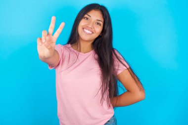  Kadın parmaklarını kameraya doğrultuyor ve birini seçiyor. Seni tavsiye ederim. En iyi seçenek. Pembe tişört giyen esmer, güzel bir kadın mavi arka plana poz veriyor.