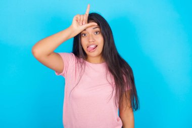  Alnında parmak olan bir kadın jesti eziklerin jestini yapar insanların dil göstermesiyle dalga geçer. Pembe tişört giyen esmer, güzel bir kadın mavi arka plana poz veriyor.