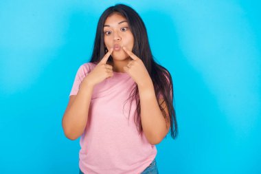  Kadınlar şaşı bakarlar ve balık dudaklarını komik bir şekilde buruştururlar. Pembe tişört giyen esmer, güzel bir kadın mavi arka plana poz veriyor.