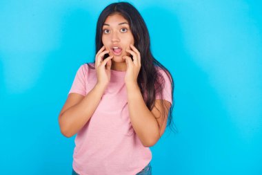 Dilsiz kadın, kameraya şoke edici bakışlara karşı elleri açık, ağzı açık bir şekilde tepki veriyor. Pembe tişört giyen esmer, güzel bir kadın mavi arka plana poz veriyor.