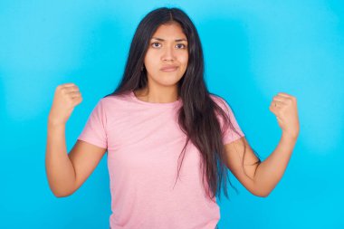Öfkeli kadın, öfkeyle yanakları üfler ve yumruklarını kaldırır. Öfke ve agresif duyguları ifade eder. Öfkeli model. Pembe tişört giyen esmer, güzel bir kadın mavi arka plana poz veriyor.