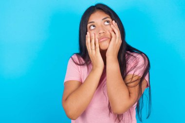 Stresli, esmer, pembe tişörtlü bir kadın mavi arka planda yüzüne dokunuyor ve çaresiz görünüyor.