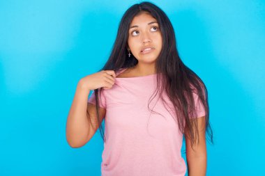 Stresli, esmer, pembe tişörtlü bir kadın mavi arka planda, sanki seksi hissediyormuş gibi boynunu çekiyor.