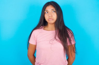 Üzgün, genç, güzel, pembe tişörtlü esmer kadın mavi arka plana bakıyor.