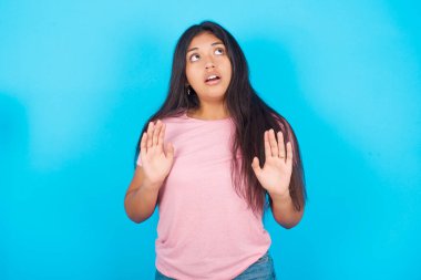 Pembe tişörtlü, esmer, korkmuş genç ve güzel bir kadın mavi arka plana yaslanmış, avuçlarını kaldırıyor ve yukarı bakıyor.