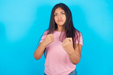 Mavi arka planda pembe tişörtlü, sinirli, genç, güzel esmer kadın yumruklarını sıkıyor ve saldırganca kameraya bakıyor.