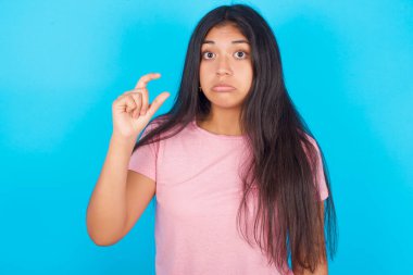 Pembe tişörtlü, hayal kırıklığına uğramış esmer bir kadın mavi arka planda parmaklarıyla küçük bedenini gösteriyor.