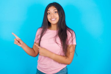 Pembe tişörtlü, mavi arka planlı, esmer, genç, güzel bir kadın. İki eliyle kameraya bakıyor.