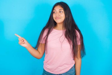 Sakin, güzel, esmer, pembe tişörtlü bir kadın mavi arka planı işaret ediyor ve yan bakıyor.