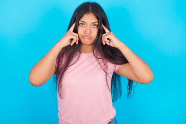 Mavi arka plana karşı pembe tişörtlü esmer kadın düşünme sürecini taklit ediyor ve işaret parmaklarını şakaklarında tutuyor.