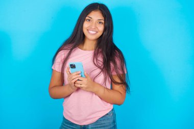 Gülümseyen, pembe tişörtlü esmer bir kadın mavi arka planda kameraya bakıyor ve iki eliyle akıllı telefonu tutuyor.