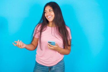 Mavi arka planda pembe tişörtlü, mutlu, genç, güzel esmer kadın bir elini ileri uzatıyor ve başka bir elde akıllı telefon tutuyor. 