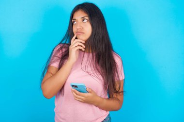 Mavi arka planda pembe tişörtlü, düşünceli, genç, güzel esmer kadın işaret parmağını dudaklarının yanında tutuyor ve başka bir elde akıllı telefon tutuyor.