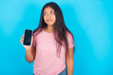 Kafası karışmış, pembe tişörtlü esmer bir kadın mavi arka plana bakıyor ve akıllı telefonunun siyah ekranını gösteriyor.