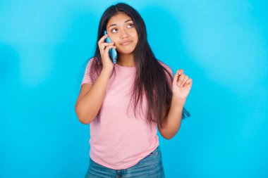 Pembe tişörtlü, mavi arkaplanlı esmer bir kadın cep telefonuyla konuşuyor.