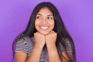 Tatmin olmuş, çizgili tişörtlü, mor arkaplanlı esmer bir kadın. Çenesini iki eliyle destekliyor ve yan bakıyor.