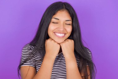 Mor arkaplanlı çizgili tişörtlü, genç ve güzel İspanyol kadın çenesini iki eliyle destekliyor ve gözlerini kapalı tutuyor.