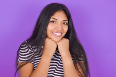 Mor arkaplanlı çizgili tişörtlü, güzel, esmer kadın çenesini iki eliyle destekliyor.